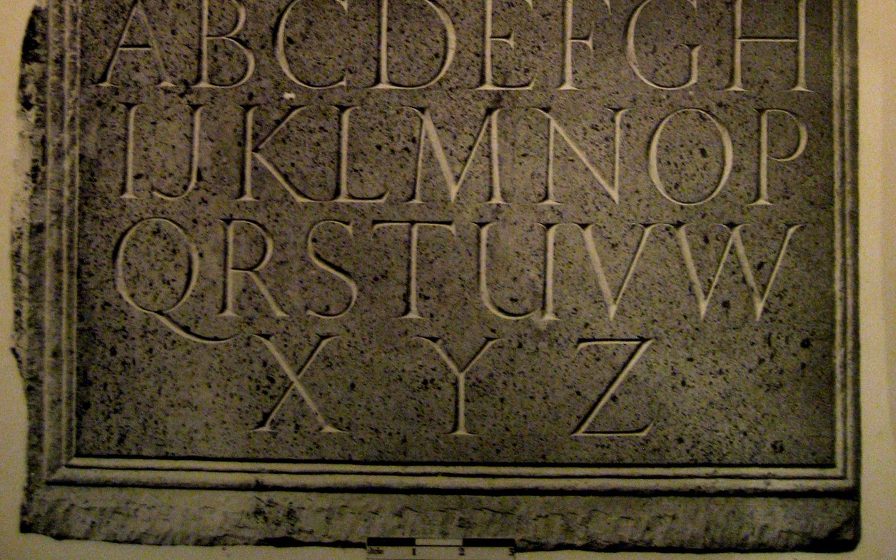 Glyphic: Roman capitals incised in stone, photographed from a printed plate, flared stems and small wedge serifs.